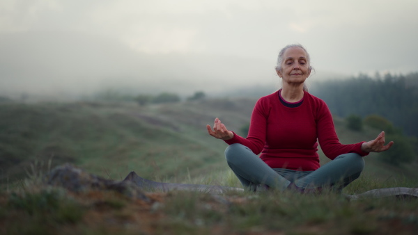 A senior woman doing breathing exercise in nature on early morning with fog and mountains in background.