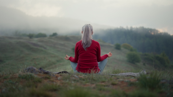 A senior woman doing breathing exercise in nature on early morning with fog and mountains in background.