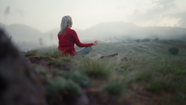 A senior woman doing breathing exercise in nature on early morning with fog and mountains in background.