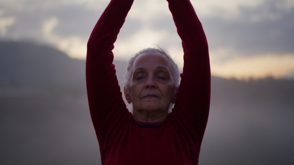 A senior woman doing breathing exercise in nature on early morning with fog and mountains in background.