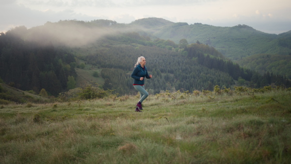 A senior woman jogging in nature on early morning with fog and mountains in background.