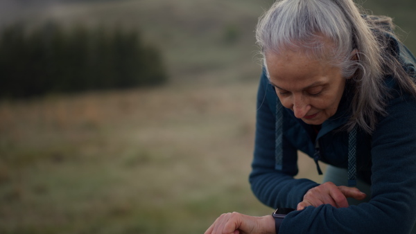 A senior woman jogger setting and looking at sports smartwatch, checking her performance in nature on early morning with fog and mountains in background.