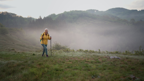 A senior woman hiking in nature on early morning with fog and mountains in background.