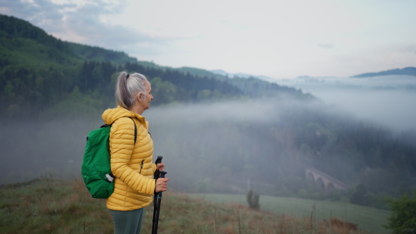 A senior woman hiking in nature on early morning with fog and mountains in background.