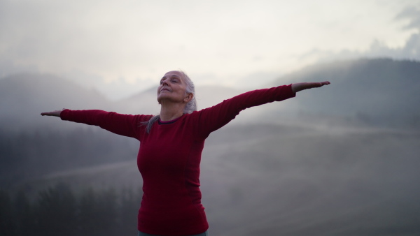 A senior woman doing breathing exercise in nature on early morning with fog and mountains in background.