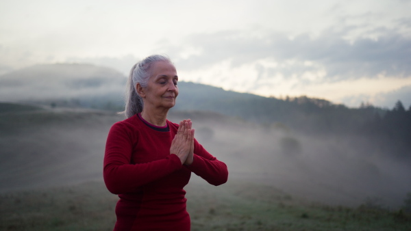 A senior woman doing breathing exercise in nature on early morning with fog and mountains in background.