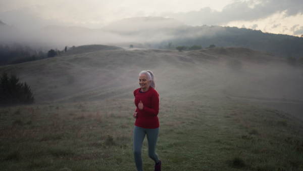 A senior woman jogging in nature on early morning with fog and mountains in background.