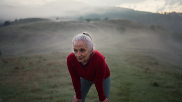 A senior woman doing breathing exercise in nature on early morning with fog and mountains in background.