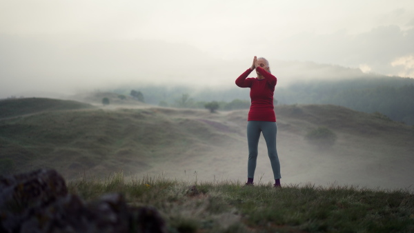 A senior woman doing breathing exercise in nature on early morning with fog and mountains in background.