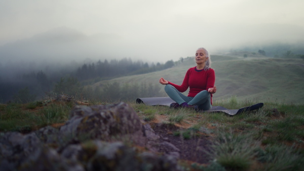 A senior woman doing breathing exercise in nature on early morning with fog and mountains in background.