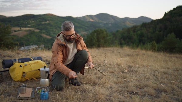 Mature male hiker in nature cutting a branch while having a coffee and snack.