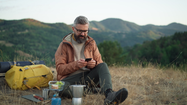 Male hiker in nature, writing message on smart phone, having a rest with coffee and sandwich.