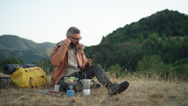 Mature male hiker sitting in nature, having a rest and listening to music on mobile pohone.