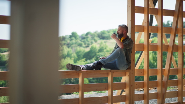 Carpenter on wooden construction site, having a rest, drinking coffee, enjoying the nature view.