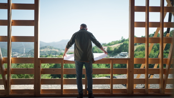 Rare view of man looking at architect drawing plans and enjoying a visionary natural view in wooden construction of eco house.