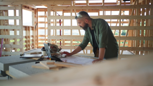 Man working on buidling site of wooden eco house, looking at plans and laptop.