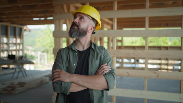 Portrait of content carpenter on building site of wooden eco house, wearing safety helmet, looking into camera, smiling.