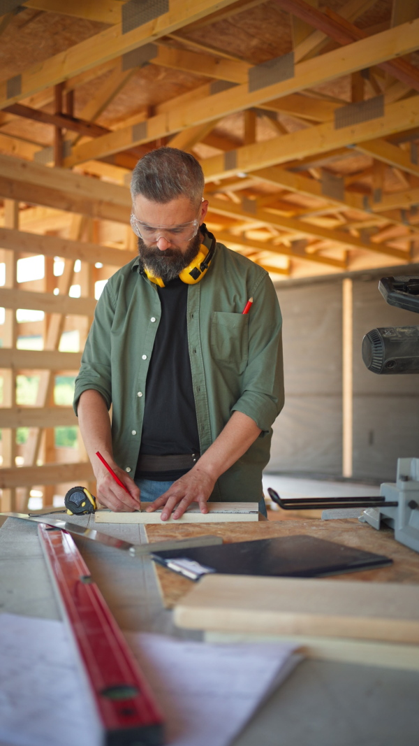 Carpenter working on buidling site of wooden eco house measuring a piece of wood. Vertical shot.