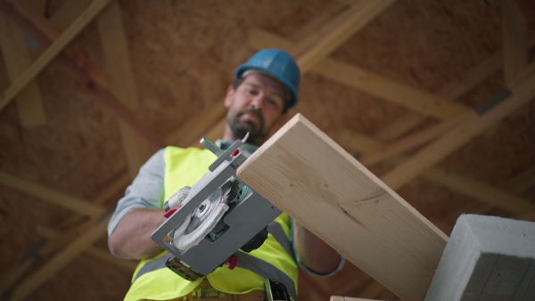 Man working on buidling site of wooden eco house cutting wood with circular drill. Low angle view.