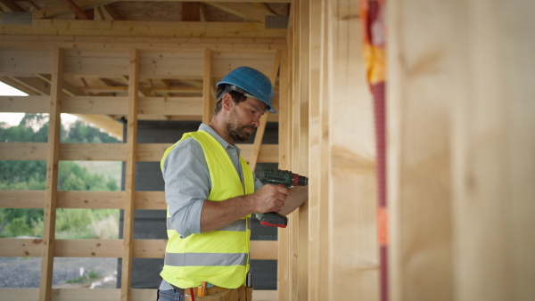 carpenter working on wooden construction site using cordless screwdriver wearinf safety vest and helmet.