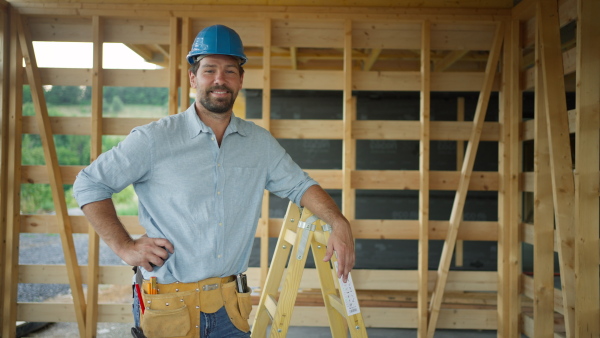 Portrait of content carpenter on building site of wooden eco house, wearing safety helmet, looking into camera, smiling.