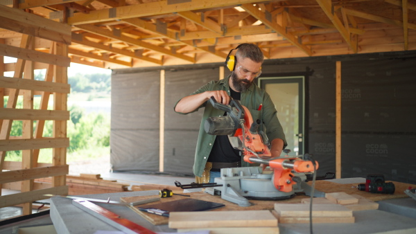 Man working on buidling site of wooden eco house cutting wood with circular drill.