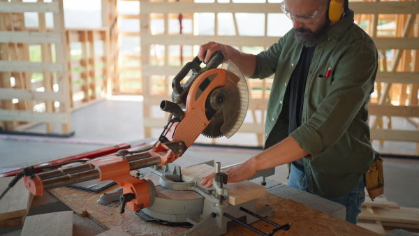 Man working on buidling site of wooden eco house cutting wood with circular drill.