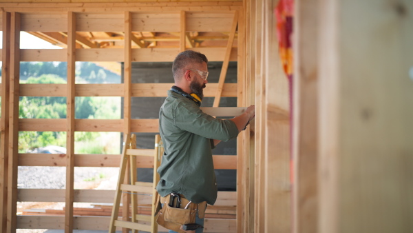 Carpenter working on buidling site of wooden eco house, measuring wooden wall.