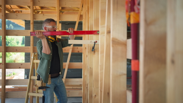 Carpenter working on buidling site of wooden eco house, measuring wooden wall using spirit level.