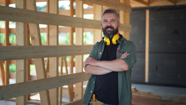 Portrait of content carpenter on building site of wooden eco house, looking into camera, smiling.