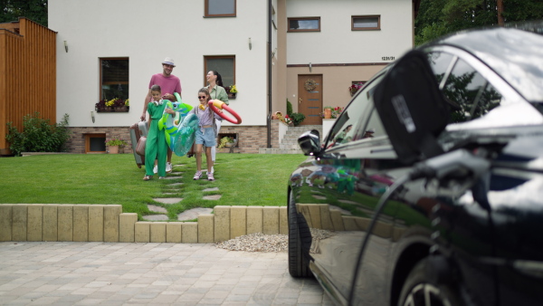 Family carrying inflatable swimming tools and luggage into electric car in driveway befoire heading off for a holiday trip.