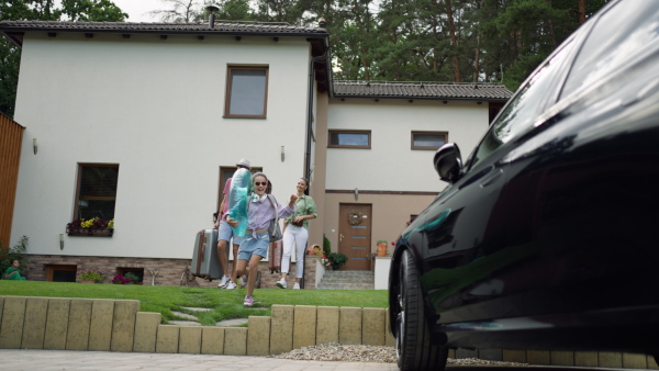 Happy and excited girl running with inflatable swimming ring to the car in driveway, heading for summer holidays. Family and house in background.