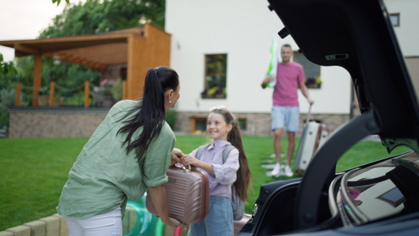 Happy family loading luggage into car before going on summer holiday. daughter handing suitcase to her mother, smiling.