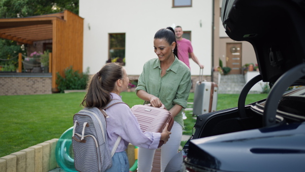 Happy family loading luggage into car before going on summer holiday. daughter handing suitcase to her mother, smiling.