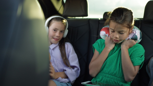 Two girls in the backseat of a car, listening to music, using digital tablet, eating an apple.