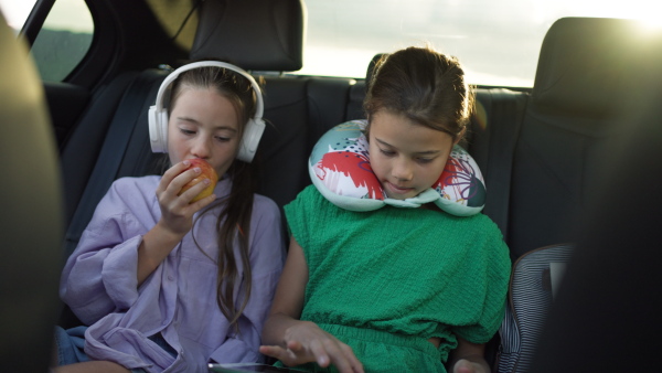 Two girls in the backseat of a car, listening to music, using digital tablet, eating an apple.