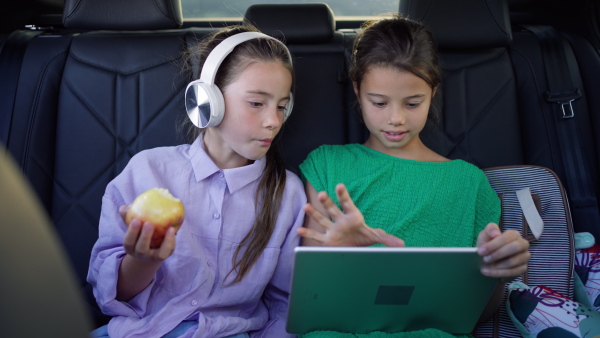 Two girls in the backseat of a car, listening to music, using digital tablet, eating an apple.