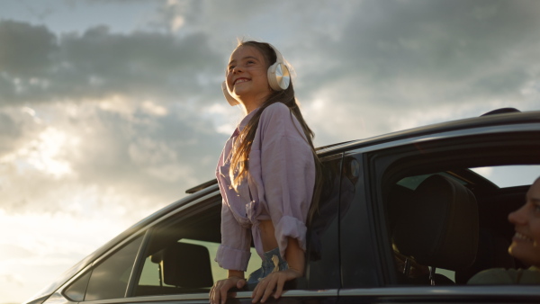 Happy girl and leaning out of car window driving at sunset, listening to music on headphones, enjoying the breeze.