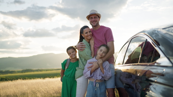 Happy family standing next to car with sunlit field in background, looking into camera.