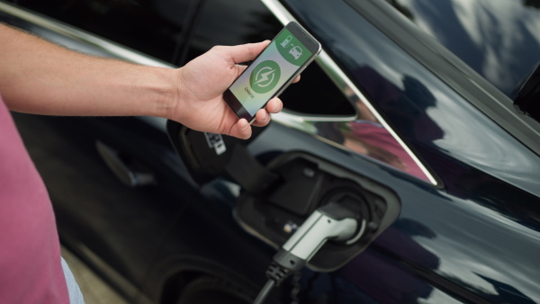 Close up of man checking mobile application on phone while charging battery of electric car. After successfull charging closing the lid.