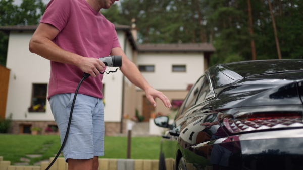 Man charging electric car in driveway. House in background. Midsection shot.