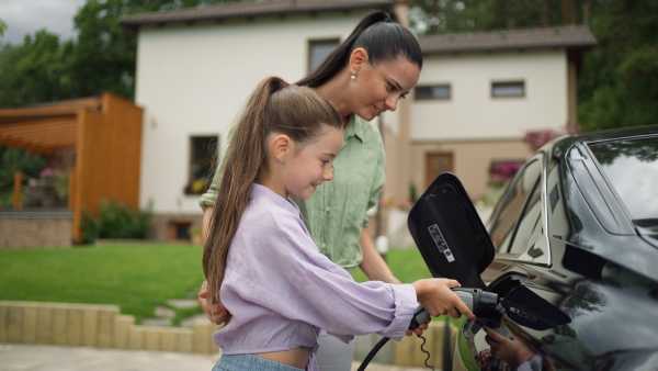 Mother and daughter charging electric car in driveway, smiling. House in background.