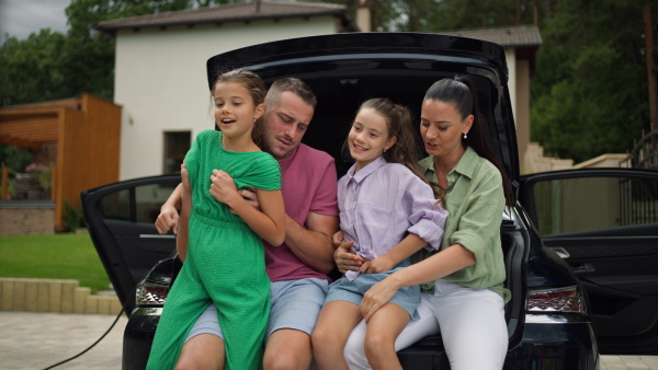 Family sitting in open electric car trunk, looking at camera, smiling.