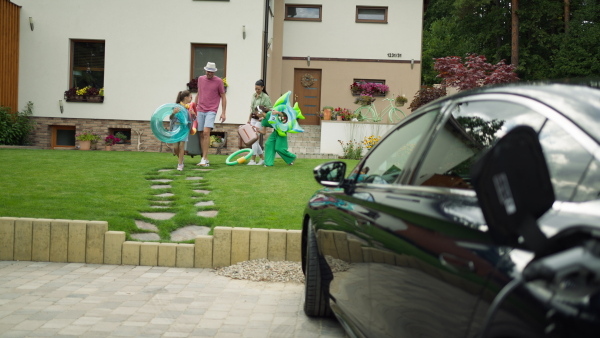 Happy family carrying inflatable swimming tools and baggage into electric car in driveway before going on holiday.