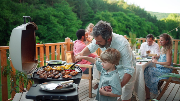 A father with little son grilling ribs and vegetable on grill during family summer garden party.