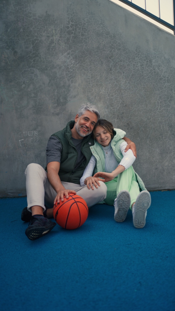 A happy father and teen daughter sitting, embracing and looking at camera outside at basketball court.