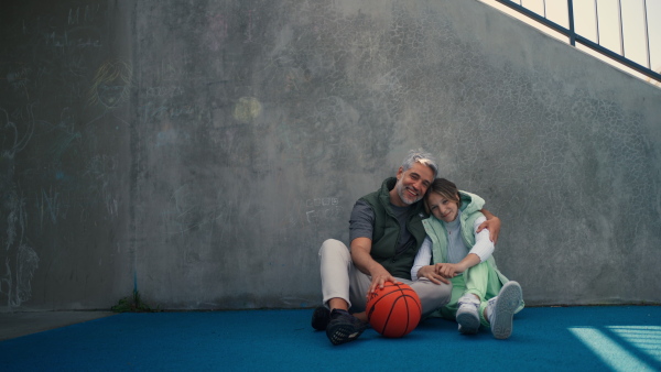 A happy father and teen daughter sitting, embracing and looking at camera outside at basketball court.