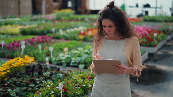 A woman in apron, employee of garden center, taking care of plants and doing check up at garden centre greenhouse.