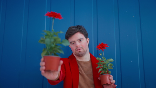 A happy young man with Down syndrome looking at camera and holding pot flowers against blue background.