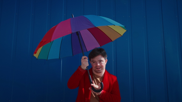 A happy young man with Down syndrome looking at camera and holding colorful umbrella against blue background.